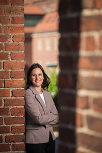 A smiling woman in a light brown jacket is leaning against a brick wall.
