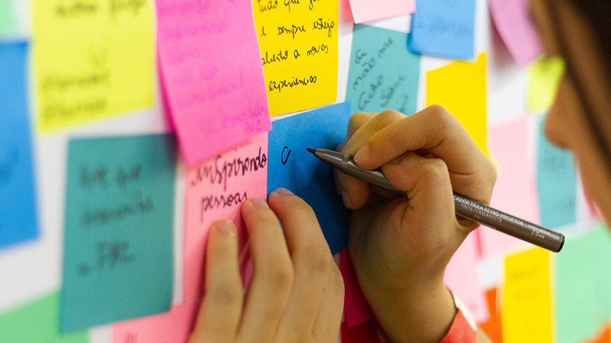 A person is writing on a post-it in front of a wall is covered with post-its in different colors.