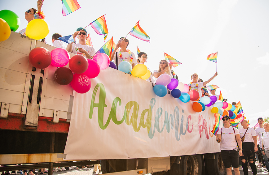 Kvinnor och män håller i pride flaggor på ett lastbilsflak som är dekorerad med färgglada ballonger och en banderoll med texten Academic Priide.