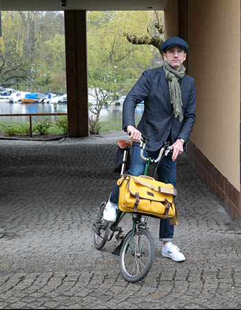 A man with a yellow bag standing by a bicycle. In the background a water canal with boats. 