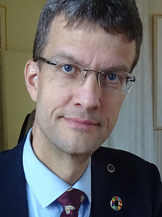 Porträttfoto: En man med kort mörk frisyr, glasögon, slips och kavaj.