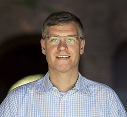 A man with dark grey hair, glasses and a white shirt.