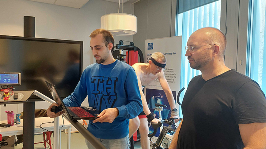 Två män med en dator. Man på träningscykel i bakgrunden. 