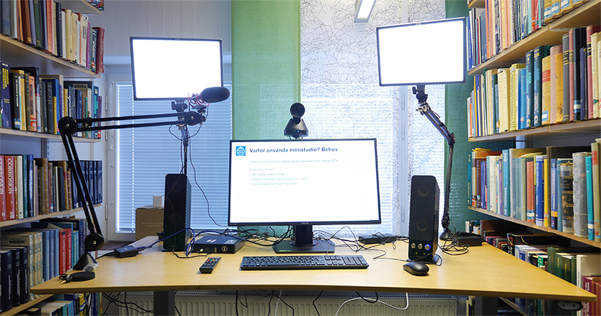 Foto på nspelningsutrustning: skärm, tangentbord, 2 högtalare och lampor på bord mellan bokhyllor.