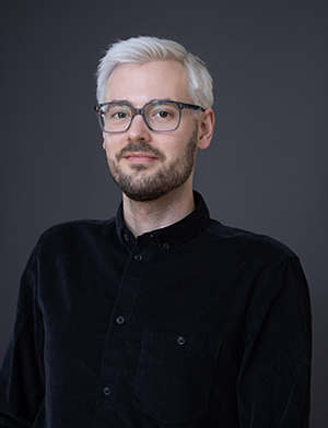 Portrait photo: A man wearing a black shirt and glasses, standing in front of a dark background.