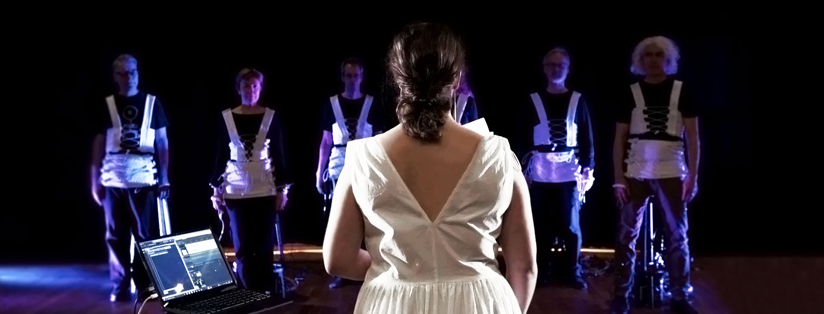 A female opera singer stands in front of a group of six people dressed with the Corsetto garment.