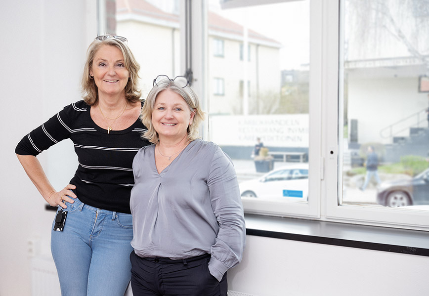 Two women standing in a room in front of a big window.