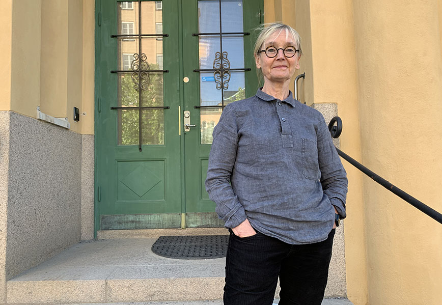 Porträttfoto: En blond kvinna med glassögon utanför en gul byggnad med grön port.