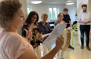 Four people stand in a room with paper in their hands. One of the women is holding a tambourine.