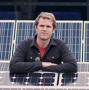 Portrait photo: A young blond man wearing a dark jacket sitting among solar panels. Photo: KTH.