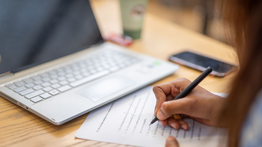 person writing notes with paper and pencil next to their computer