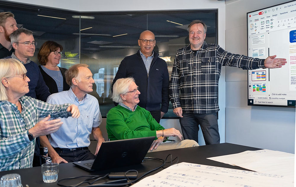 En grupp människor vid ett konferensbord där flertalet är glada och en person pekar på en stor skärm