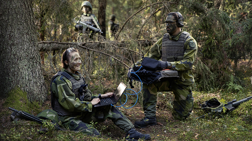 Two soldiers in camouflage in the forest. A soldier sits on the ground with a laptop in their lap.