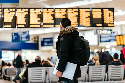 A man with a bag and a laptop looking at a time table board in a big hall.