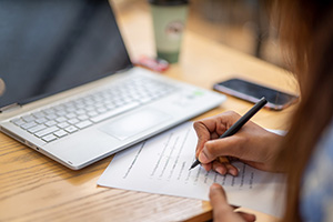 A hand writing on a paper in front of a computer. 