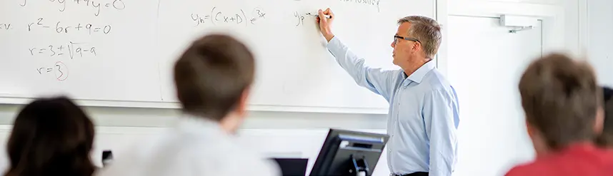 A teacher stands at a whiteboard and writes. Students are visible in the foreground.