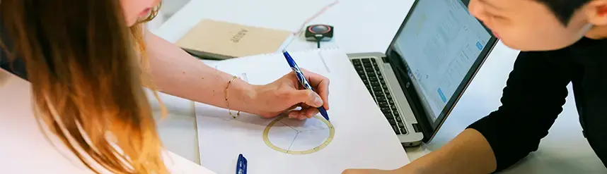 Two people sit by a paper and a laptop. One of them is drawing a circle.