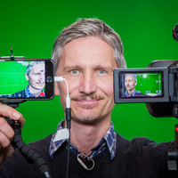 Syntolkning: Jonas Thorén mot grön bakgrund, håller en videokamera och en smartphone framför sig. 