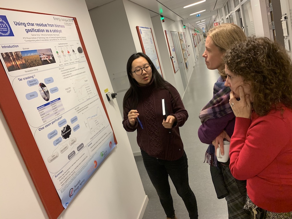 In the corridor of the Department of Chemical Engineering: a woman presents a poster on the wall explaining the use of carbon dioxide from biomass gasification as a catalyst for two women from the Energy Platform who are visiting.