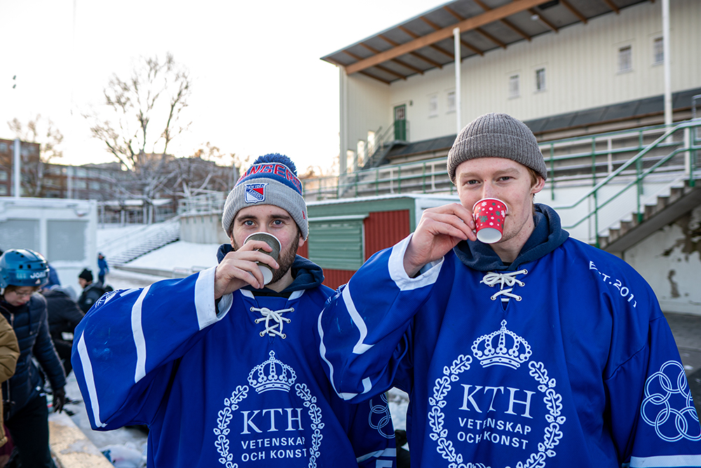 Anton Haga Lööf and Oskar Ringström at Östermalm IP. They are wearing hockey equipment with the KTH logo on and they drink something from mugs.
